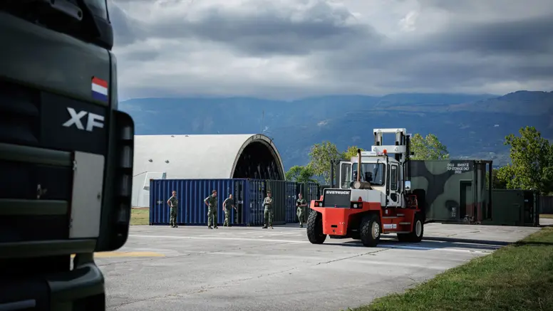 Een heftruck met een container rijdt over een betonnen ondergrond tussen gras gelegen. Links meerdere containers en een oude vliegtuigshelter. Op de achtergrond bergen.
