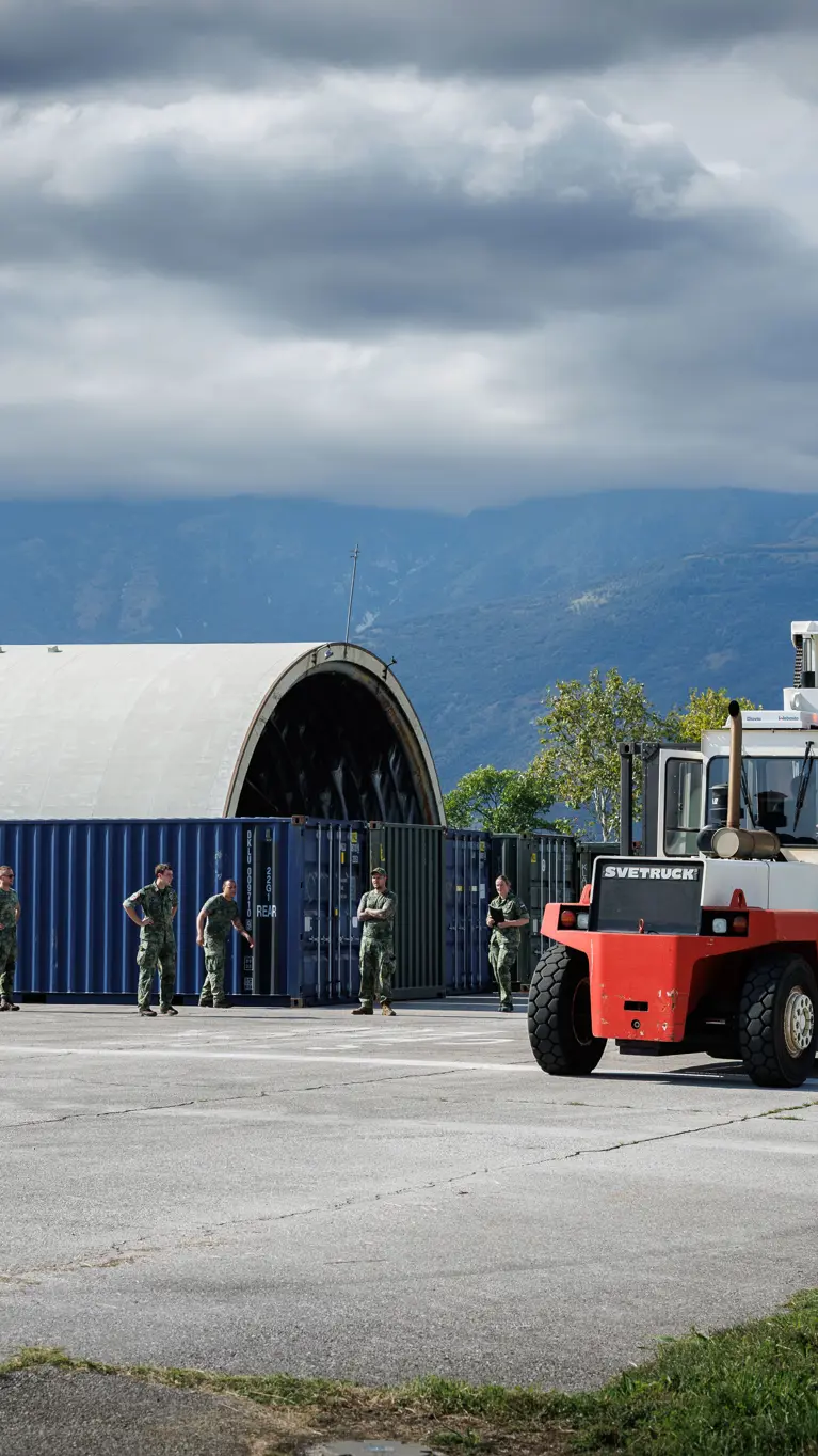 Een heftruck met een container rijdt over een betonnen ondergrond tussen gras gelegen. Links meerdere containers en een oude vliegtuigshelter. Op de achtergrond bergen.
