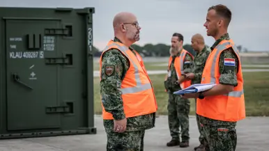 Twee militairen in oranje hesjes staan buiten tegenover elkaar. Een van heen heeft een mapje met papieren in zijn hand en kijkt op zijn horloge. Op de achtergrond staan nog twee militairen, een van hen draagt ook een oranje hesje. Naast hen een groene container.