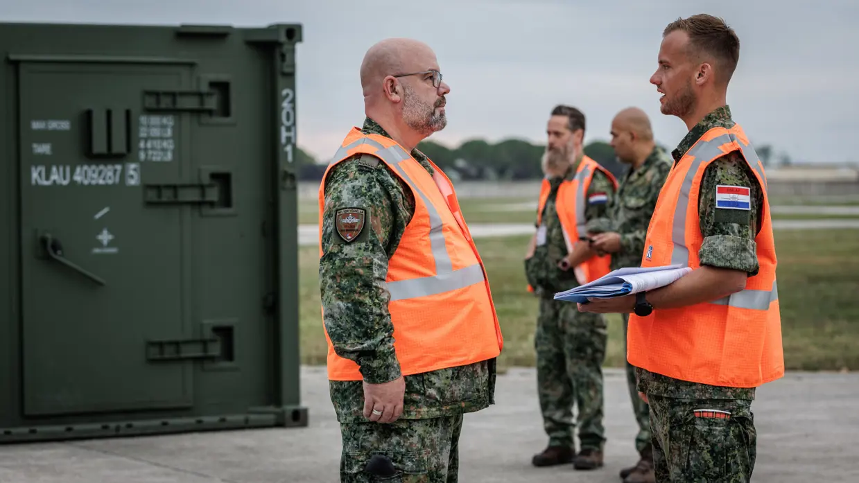 Twee militairen in oranje hesjes staan buiten tegenover elkaar. Een van heen heeft een mapje met papieren in zijn hand en kijkt op zijn horloge. Op de achtergrond staan nog twee militairen, een van hen draagt ook een oranje hesje. Naast hen een groene container.