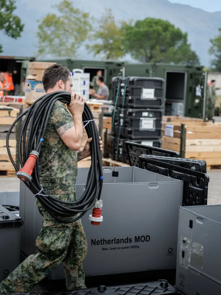 Een militair loopt met een stroomkabelbundel over het terrein tussen de boxen, pallets en containers. Op een grijze box staat de tekst ‘Netherlands MOD’. Op de achtergrond enkele bomen en een bergketen.