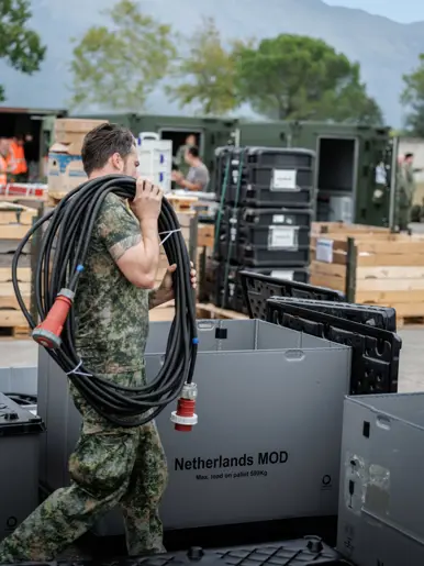 Een militair loopt met een stroomkabelbundel over het terrein tussen de boxen, pallets en containers. Op een grijze box staat de tekst ‘Netherlands MOD’. Op de achtergrond enkele bomen en een bergketen.