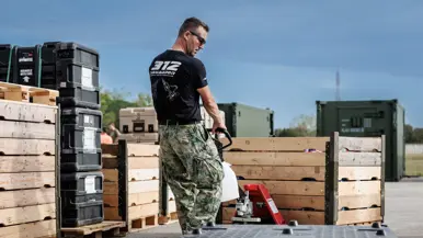 Een militair verplaatst met een steekwagen een palletbox. Om hem heen staan nog meer palletboxen en dozen. Op de achtergrond staan groene containers. Hij draagt een zwart T-shirt met daarop de tekst ‘312 Squadron’ met daaronder een grafische weergave van twee F-35’s.