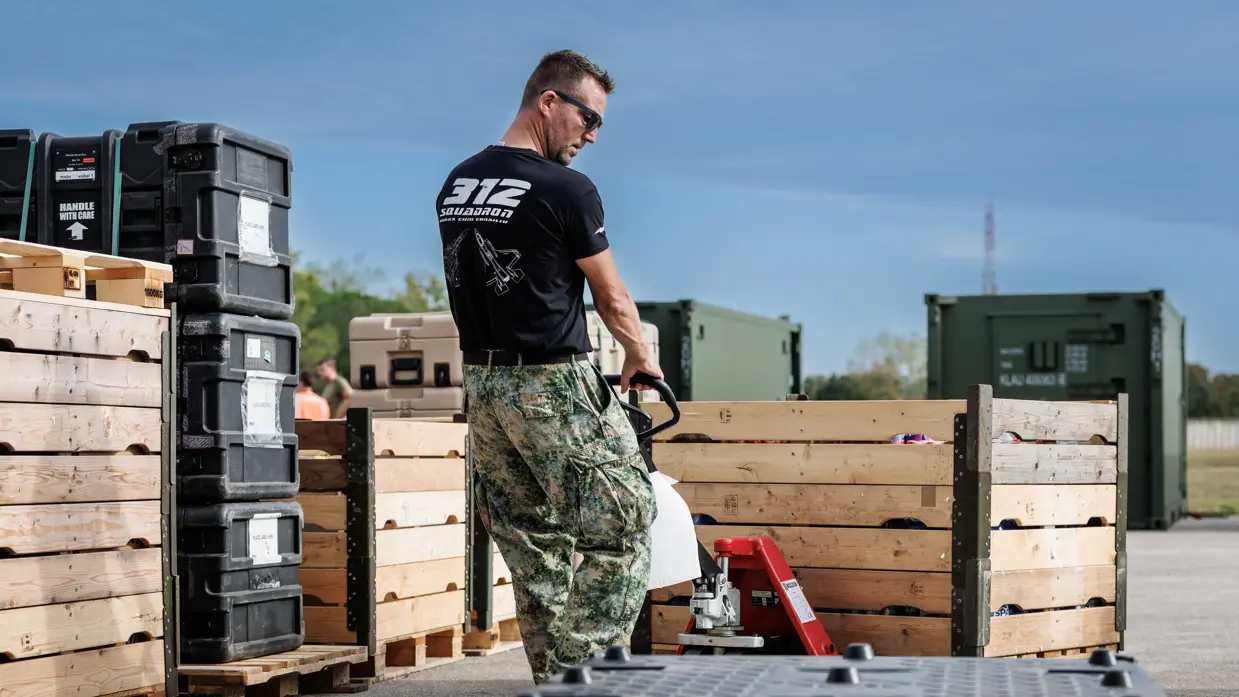 Een militair verplaatst met een steekwagen een palletbox. Om hem heen staan nog meer palletboxen en dozen. Op de achtergrond staan groene containers. Hij draagt een zwart T-shirt met daarop de tekst ‘312 Squadron’ met daaronder een grafische weergave van twee F-35’s.