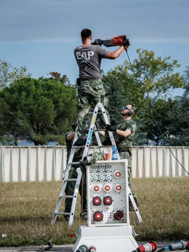 Drie militairen aan het werk bij een dubbele ladder, twee van hen houden de lader vast, een staat erop. Naast een groepenkast met op de grond een aantal stroomkabels.