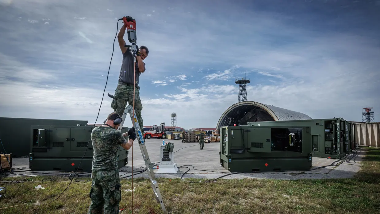 Twee militairen aan het werk bij een dubbele ladder, een houdt de lader vast, de ander staat erop. Achter hen een groepenkast, twee grote groene stroomgeneratoren en een aantal stroomkabels die over het terrein achter groene containers liggen. Een oude shelter op de achtergrond.
