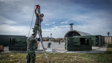 Twee militairen aan het werk bij een dubbele ladder, een houdt de lader vast, de ander staat erop. Achter hen een groepenkast, twee grote groene stroomgeneratoren en een aantal stroomkabels die over het terrein achter groene containers liggen. Een oude shelter op de achtergrond.