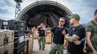 Twee militairen met zwarte shirts in gesprek tussen de werkende andere militairen. Om hen heen staan palletboxen en dozen. Op de achtergrond een vliegtuigshelter.