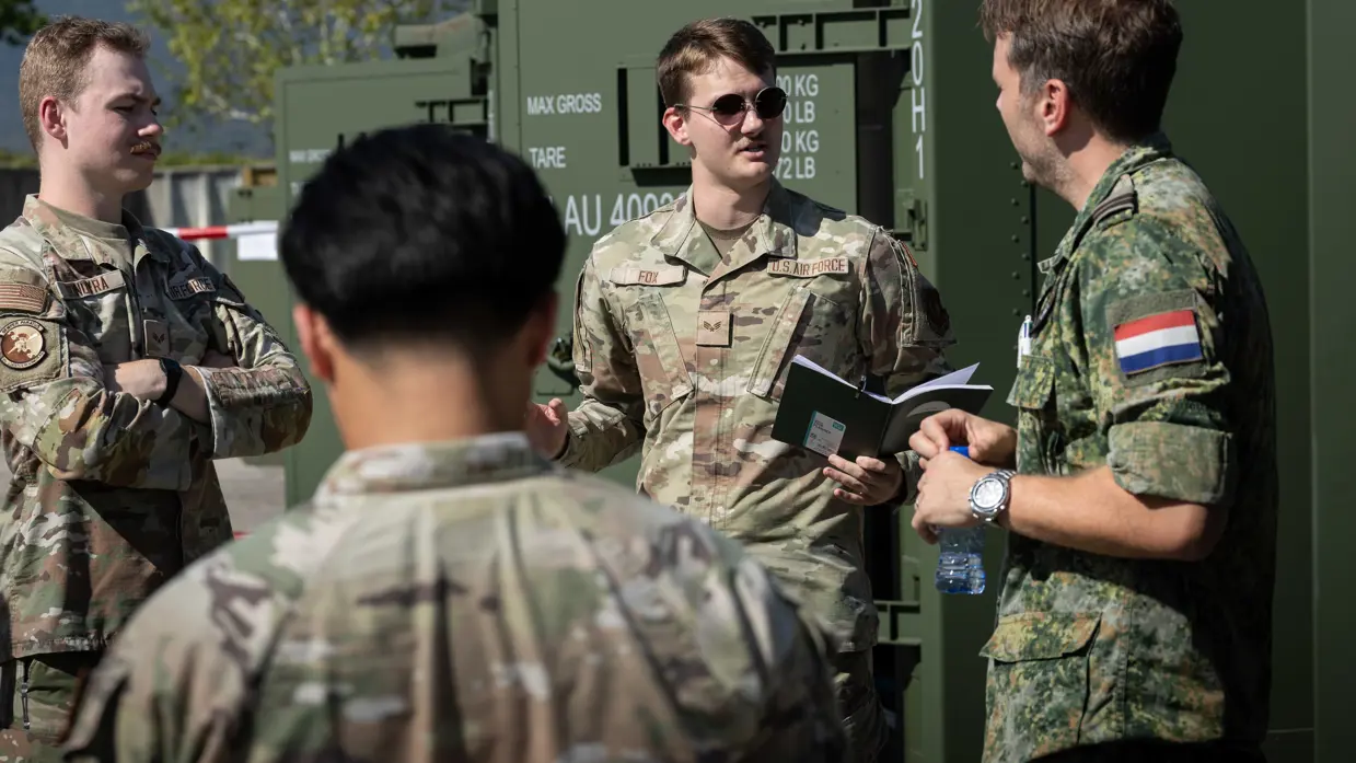 De detachementscommandant in een kringgesprek met drie Amerikaanse militairen, buiten naast een rij groene containers. Een van de militairen heeft een klein notitieboekje in de hand.  