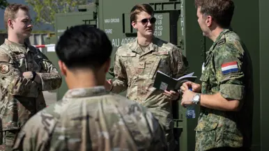 De detachementscommandant in een kringgesprek met drie Amerikaanse militairen, buiten naast een rij groene containers. Een van de militairen heeft een klein notitieboekje in de hand.  