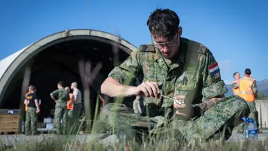 Militair in kleermakerszit in het gras met een stekker, een stroomkabel en schroevendraaier. Op de achtergrond twee groepjes militairen staand in overleg op het kamp in opbouw.