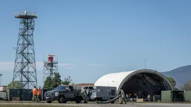Buitenterrein in opbouw met onder meer twee vaste masten en een oude vliegtuigshelter. Militairen zijn onder andere in de weer met een grijze aanhanger waaruit buizen komen. Deze staat achter een voertuig. Twee militairen met oranje hesjes kijken toe. Op de achtergrond een deel van een bergketen.