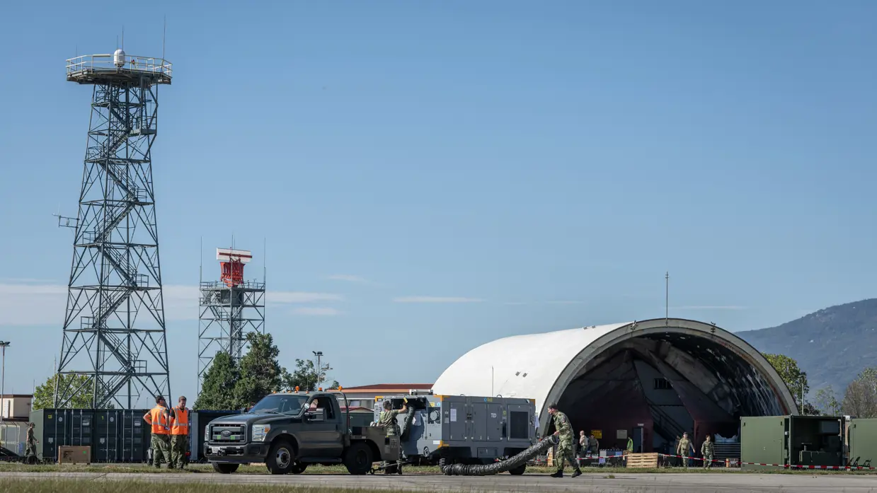 Buitenterrein in opbouw met onder meer twee vaste masten en een oude vliegtuigshelter. Militairen zijn onder andere in de weer met een grijze aanhanger waaruit buizen komen. Deze staat achter een voertuig. Twee militairen met oranje hesjes kijken toe. Op de achtergrond een deel van een bergketen.