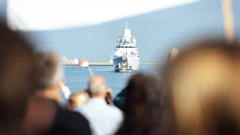 Een marineschip komt aan in de haven terwijl familie aan de kade staat te wachten.