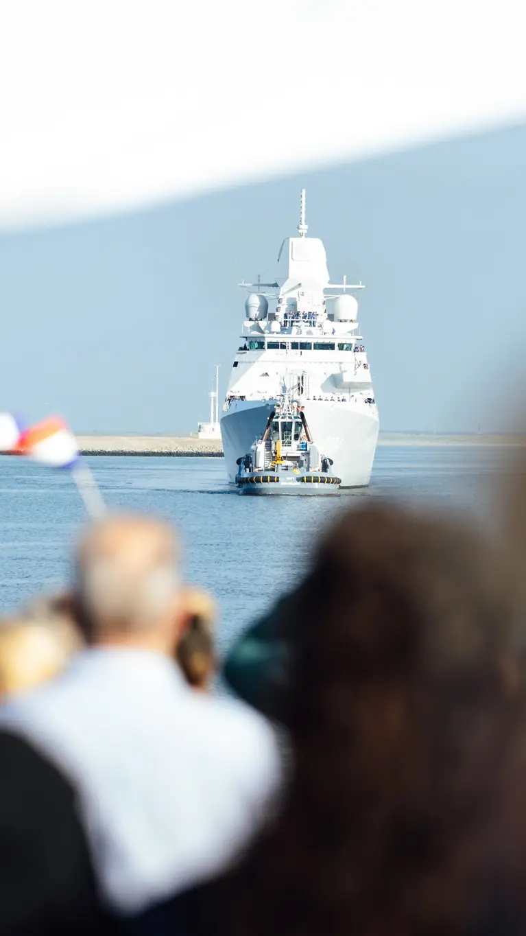 Een marineschip komt aan in de haven terwijl familie aan de kade staat te wachten.