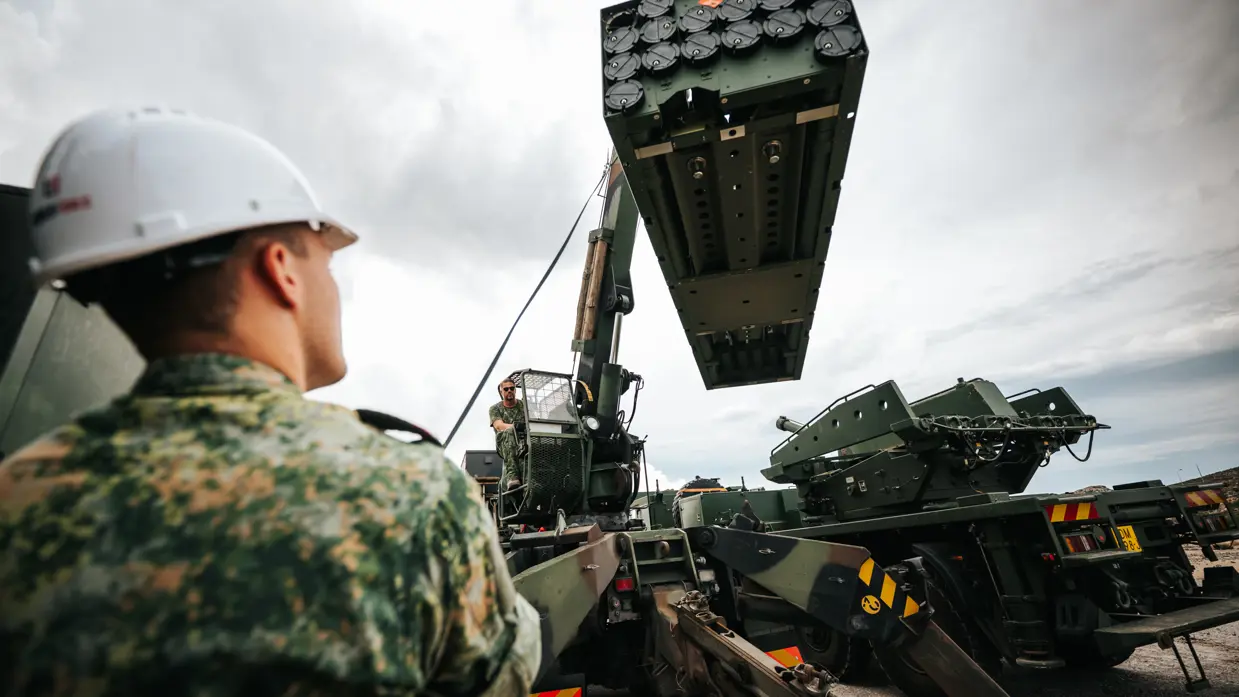 Een militair met witte helm op kijkt hoe een kraanmachinist een serie raketten hijst.