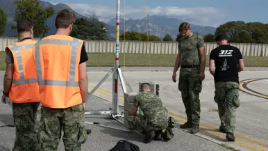 Drie militairen aan het werk aan een stalen constructie, een van hen draagt een zwart T-shirt met daarop de tekst ‘312 Squadron’ met daaronder een grafische weergave van een F-35. Twee militairen in oranje hesjes kijken toe. Op de achtergrond een bergketen.