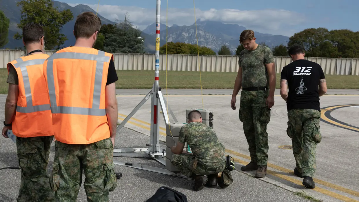 Drie militairen aan het werk aan een stalen constructie, een van hen draagt een zwart T-shirt met daarop de tekst ‘312 Squadron’ met daaronder een grafische weergave van een F-35. Twee militairen in oranje hesjes kijken toe. Op de achtergrond een bergketen.