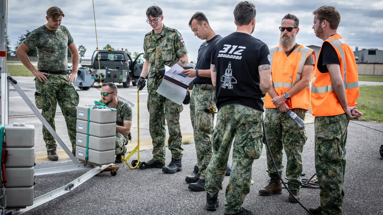 Vier militairen aan het werk aan een stalen constructie, een van hen heeft een mapje met papieren vast. Ernaast staan drie militairen in gesprek. Een van hen draagt een zwart T-shirt met daarop de tekst ‘312 Squadron’ met daaronder een grafische weergave van een F-35. De andere twee militairen dragen oranje hesjes.