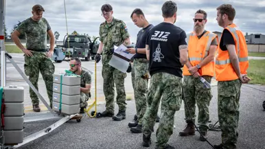 Vier militairen aan het werk aan een stalen constructie, een van hen heeft een mapje met papieren vast. Ernaast staan drie militairen in gesprek. Een van hen draagt een zwart T-shirt met daarop de tekst ‘312 Squadron’ met daaronder een grafische weergave van een F-35. De andere twee militairen dragen oranje hesjes.