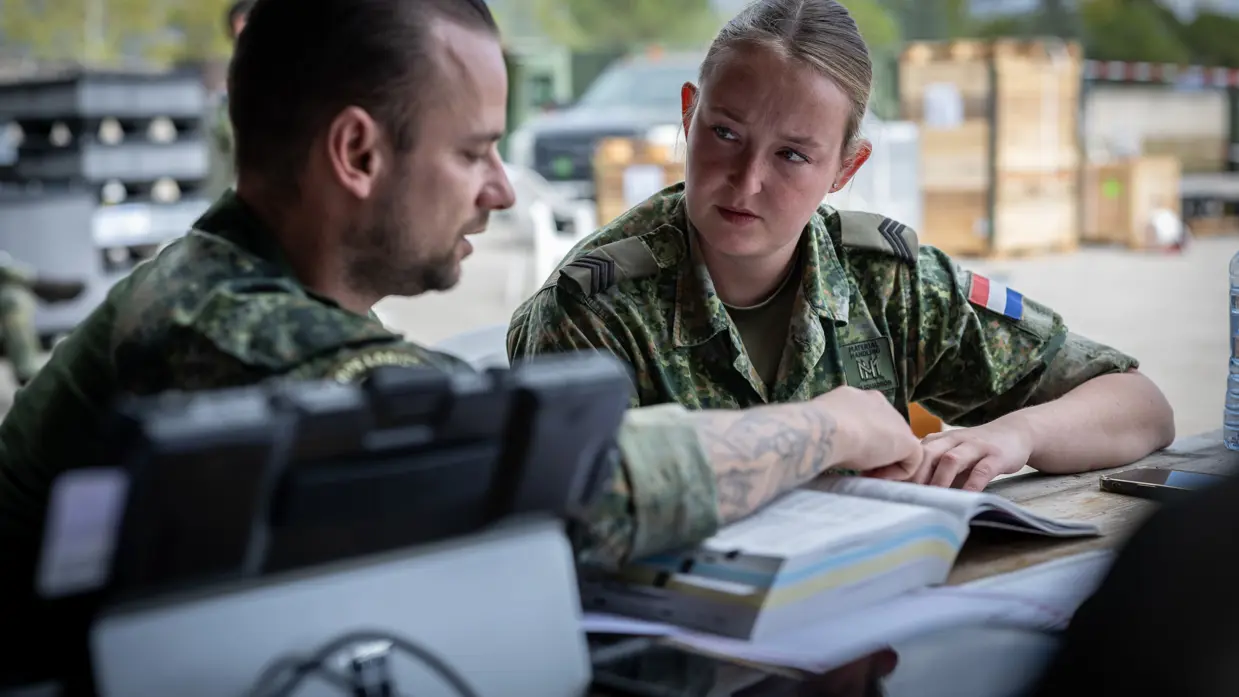 Een mannelijke en vrouwelijke militair overleggen aan een houten tafel. Ze hebben een opengeslagen boek van zo’n 8 cm voor zich en een opengeklapte laptop. Op de achtergrond op het buitenterrein staat een voertuig en meerdere houten kisten, deels gestapeld.