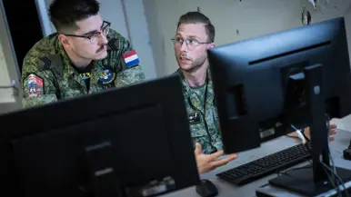 Een militair aan een bureau met twee beeldschermen overlegt met een militair die voorover gebukt naast hem staat.