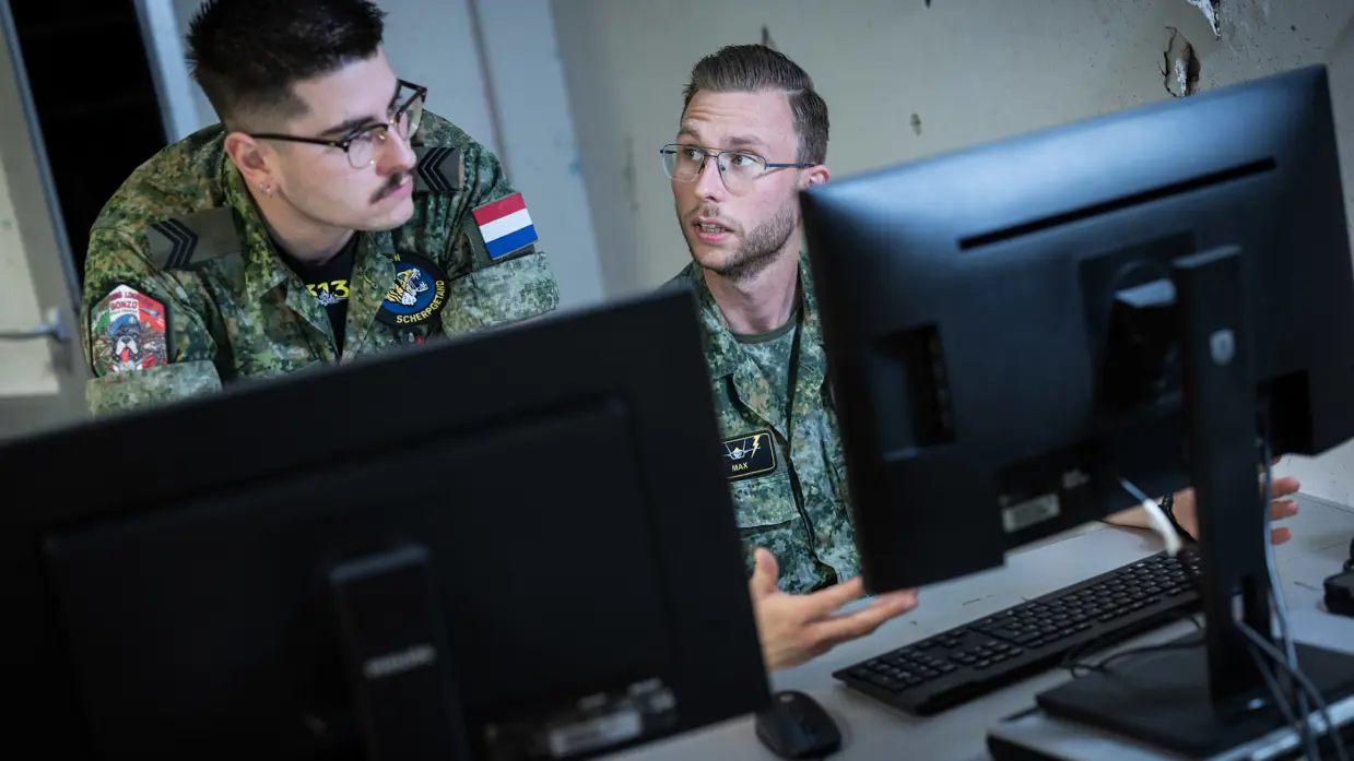 Een militair aan een bureau met twee beeldschermen overlegt met een militair die voorover gebukt naast hem staat.