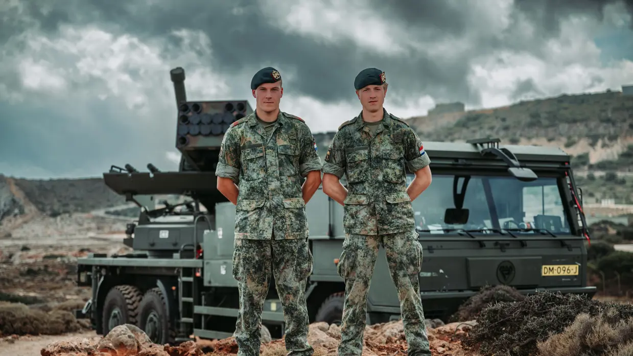 Brian en Tim staan in camouflagepak met hun handen op de rug. Op de achtergrond staat een PULS-lanceerinstallatie.