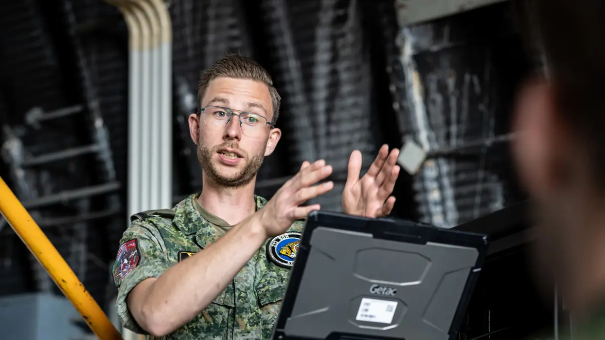 Tweede luitenant Max staat bij een systeemkast met kabels waarop hij met een ‘robuuste’ laptop aan het werk is. Op de voorgrond het silhouet van een militair waarmee hij in gesprek is.