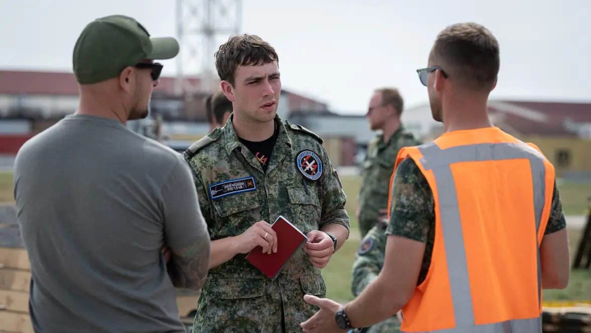 Tweede luitenant Andor met een notitieboekje in zijn hand praat in het buitenterrein met twee andere militairen. Een van hen draagt een oranje hesje.