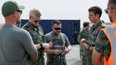 Een militair buiten in gesprek met drie militairen, twee van hen schrijven iets op in hun notitieboekje. Een militair in oranje hesje staat ernaast. Op de achtergrond een blauwe container.