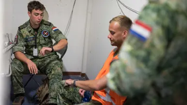 Twee militairen binnen op een klapstoel tegen een muur met kogelgaten. De rechter draagt een oranje hesje. Ze kijken beiden al pratend op hun horloge.