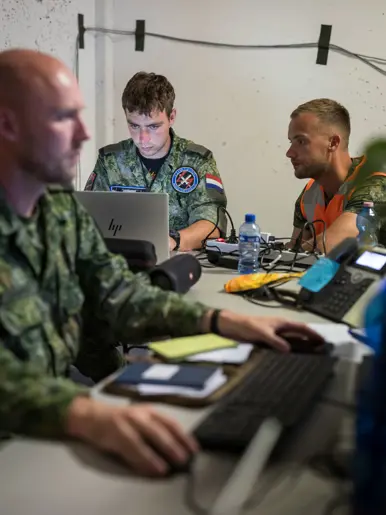 Twee militairen haaks op elkaar aan een bureau aan het werk. De een op een laptop de ander op een pc. Een militair met oranje hesje kijkt mee met een van hen. Op de met kogelgaten gevulde muur op de achtergrond zijn stroomdraden met zwarte tape bevestigd.