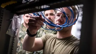 Militair met blauwe dunne kabeltjes bezig bij een systeemkast, twee militairen kijken over zijn schouder mee.