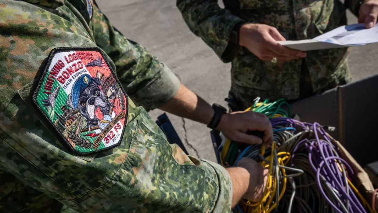 Close-up van een militair die diverse kleuren stroomkabels in een doos doet. Op zijn mouw een patch met de tekst ‘Lightning Logistics Bonzo, Italia exercise, 312 FS 2026’. Naast hem staat een militair kijkend op zijn A4-papieren in de hand.   