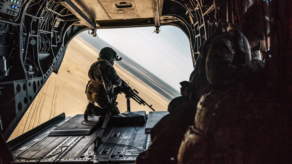 Achterin een Chinook zit een militair op de laadklep achter een mitrailleur.