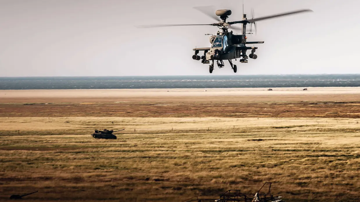 Een Apache vliegt boven een kaal bruin landschap. In de achtergrond staat een oude tank.
