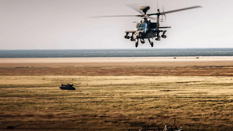Een Apache vliegt boven een kaal bruin landschap. In de achtergrond staat een oude tank.