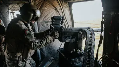 Militair in militaire helikopter, met helm en camouflage-uniform, bedient een mitrailleur bij de geopende deur. Binnenin stoelen en apparatuur zichtbaar.