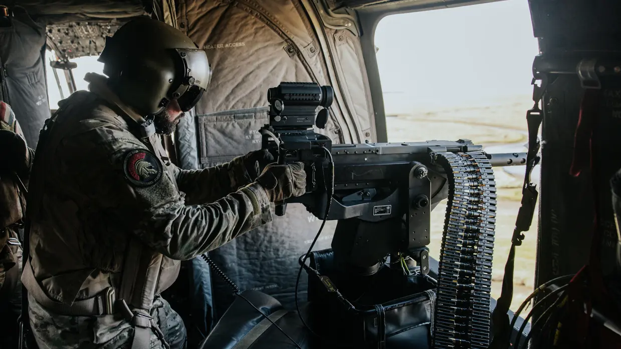 Militair in militaire helikopter, met helm en camouflage-uniform, bedient een mitrailleur bij de geopende deur. Binnenin stoelen en apparatuur zichtbaar.
