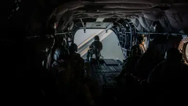 Interieur van militaire helikopter met meerdere soldaten in helm en uitrusting. Door de geopende achterklep zicht op zonovergoten kustlijn met strand en zee. Donker interieur contrasteert fel met het licht buiten.