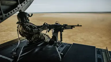 Soldaat zit in open laadklep van militaire helikopter, met helm, kogelvrij vest en camouflagekleding, achter een machinegeweer. Eronder kaal, bruin woestijnlandschap onder helderblauwe lucht.