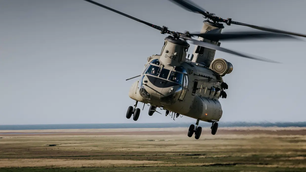Een grote militaire helikopter in volle vlucht boven een bruin landschap. 