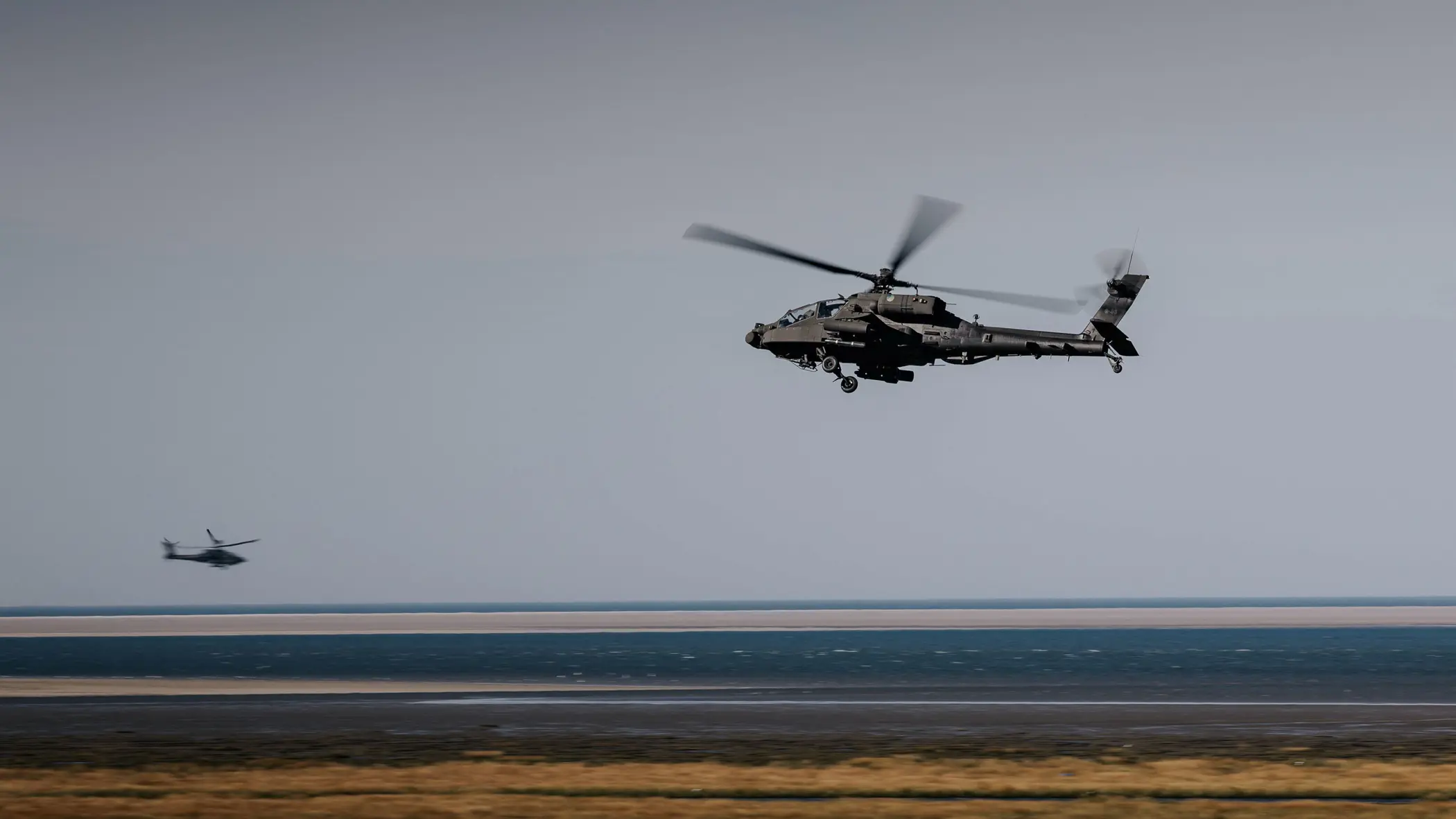Twee militaire helikopters vliegen boven een kustlandschap. Voorgrond: grote helikopter met draaiende rotoren, donkere romp. Daaronder zandstrand, blauwe zee en lichtblauwe lucht. Rustige maar dynamische sfeer.