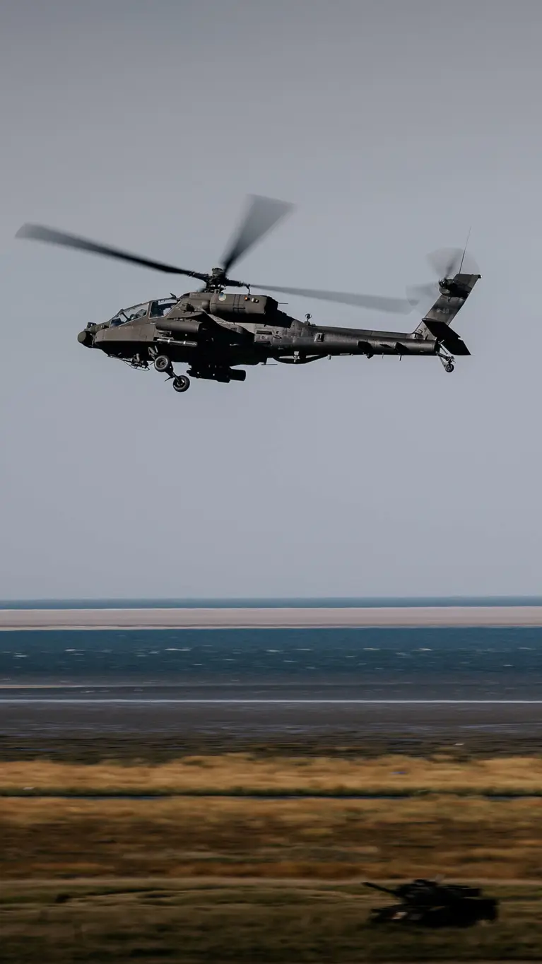 Twee militaire helikopters vliegen boven een kustlandschap. Voorgrond: grote helikopter met draaiende rotoren, donkere romp. Daaronder zandstrand, blauwe zee en lichtblauwe lucht. Rustige maar dynamische sfeer.