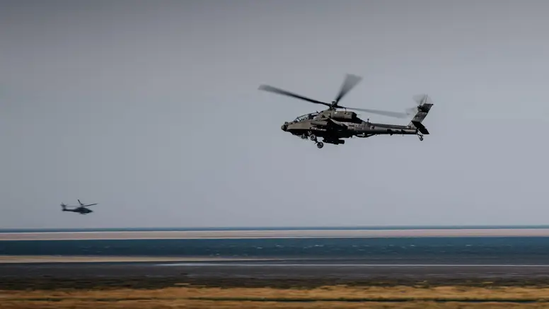 Twee militaire helikopters vliegen boven een kustlandschap. Voorgrond: grote helikopter met draaiende rotoren, donkere romp. Daaronder zandstrand, blauwe zee en lichtblauwe lucht. Rustige maar dynamische sfeer.