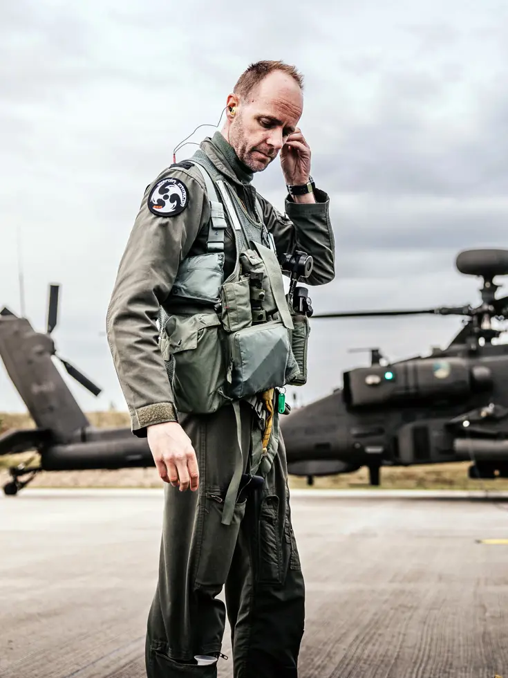Een militair loopt doet zijn oordopjes in. In de achtergrond een Apache die bewapend wordt.