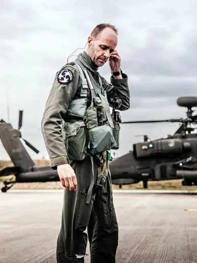 Een militair loopt doet zijn oordopjes in. In de achtergrond een Apache die bewapend wordt.