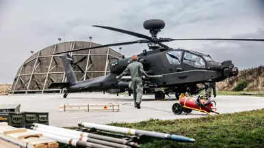 Op de voorgrond liggen raketten. Een man in overall loopt met een raket naar een Apache.