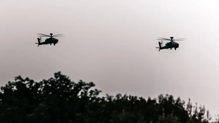 In de schemering vliegen twee Apaches met groene verlichting boven bomen.